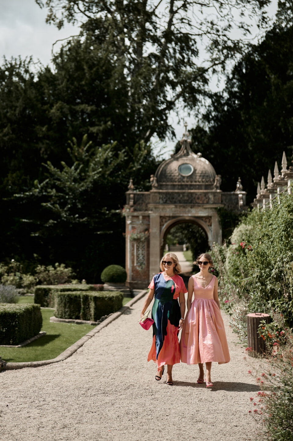 Two girls wearing pink dresses are strolling down a garden path.