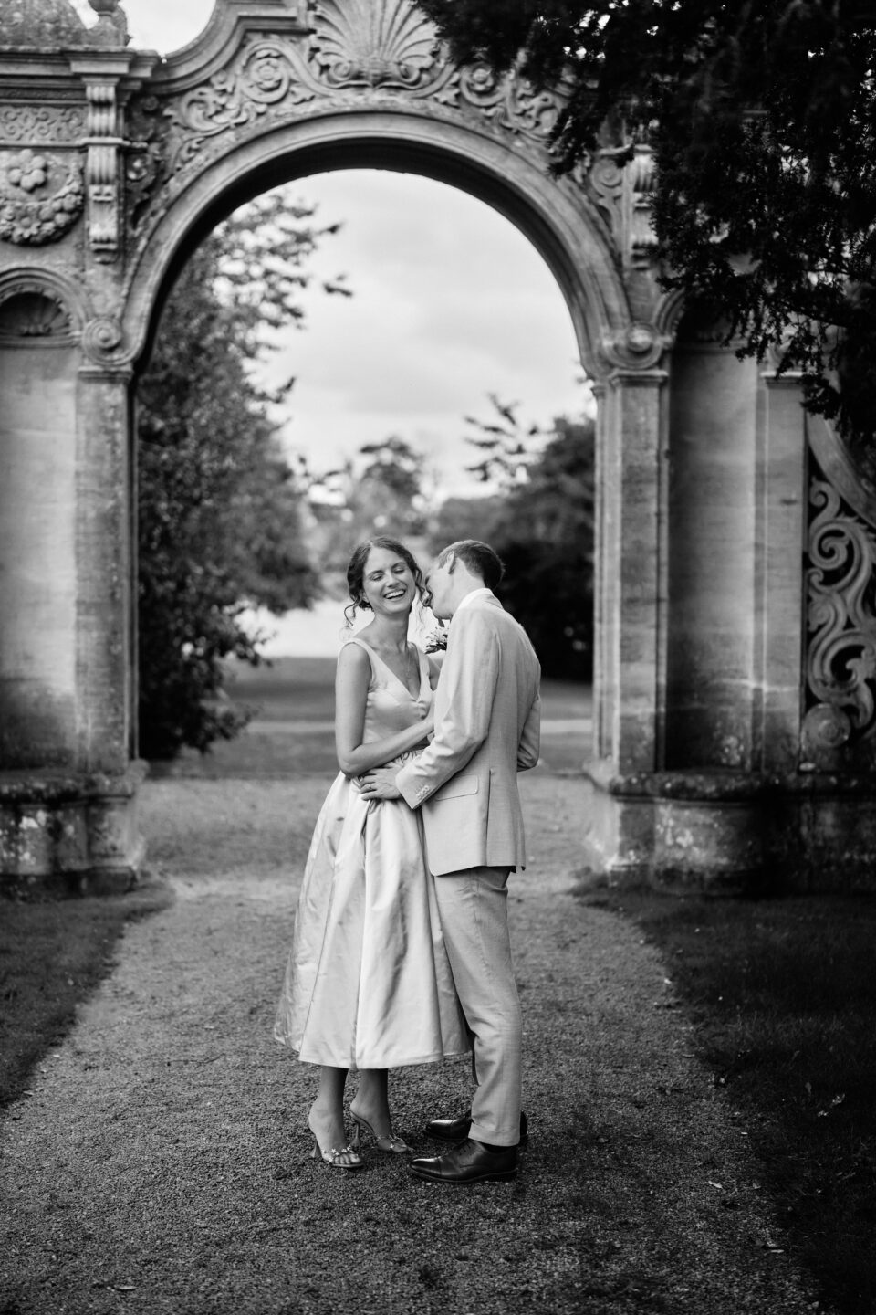 A black and white picture of a newly married couple posing in front of an arch.