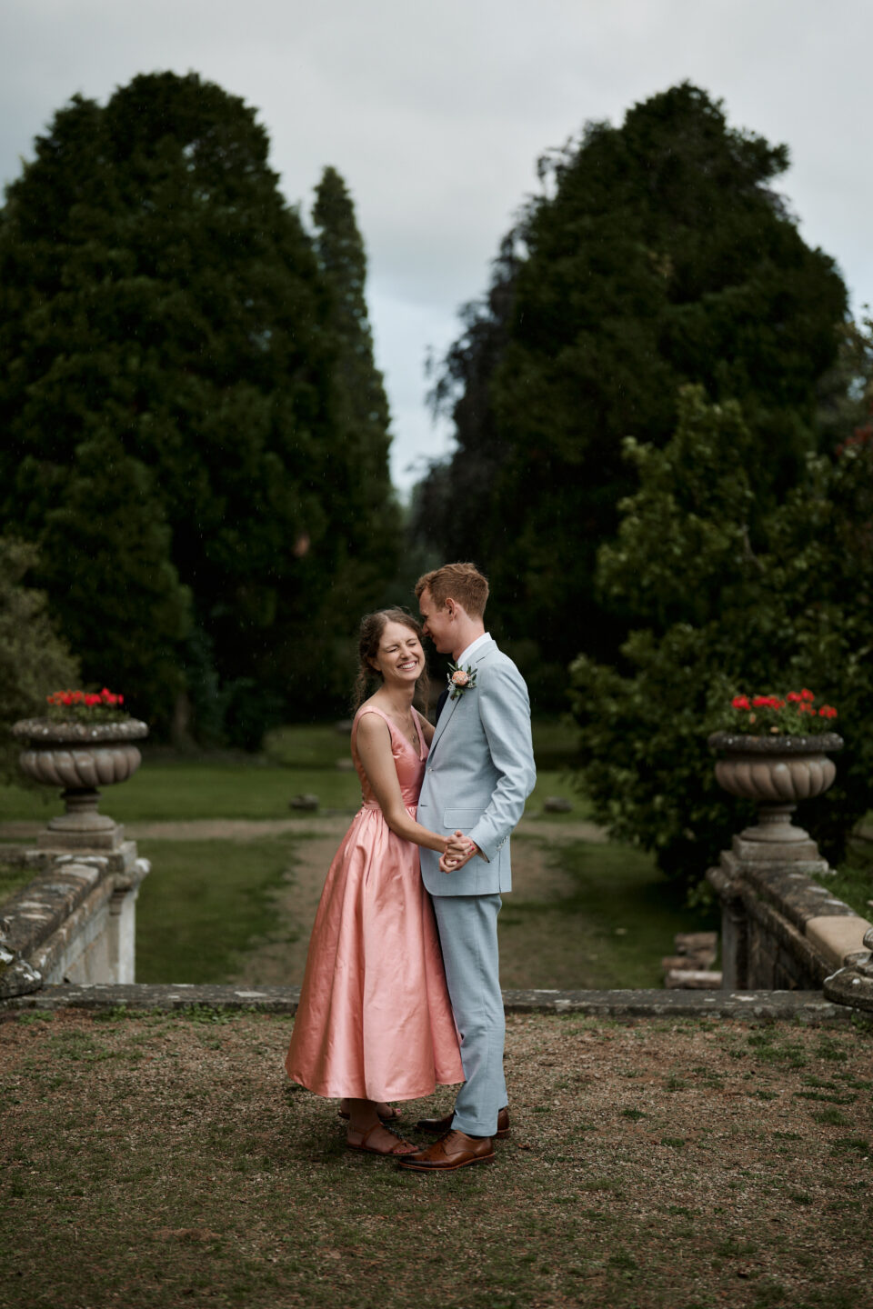 A man and woman, dressed for their wedding with the woman in a pink dress, are standing in a garden.
