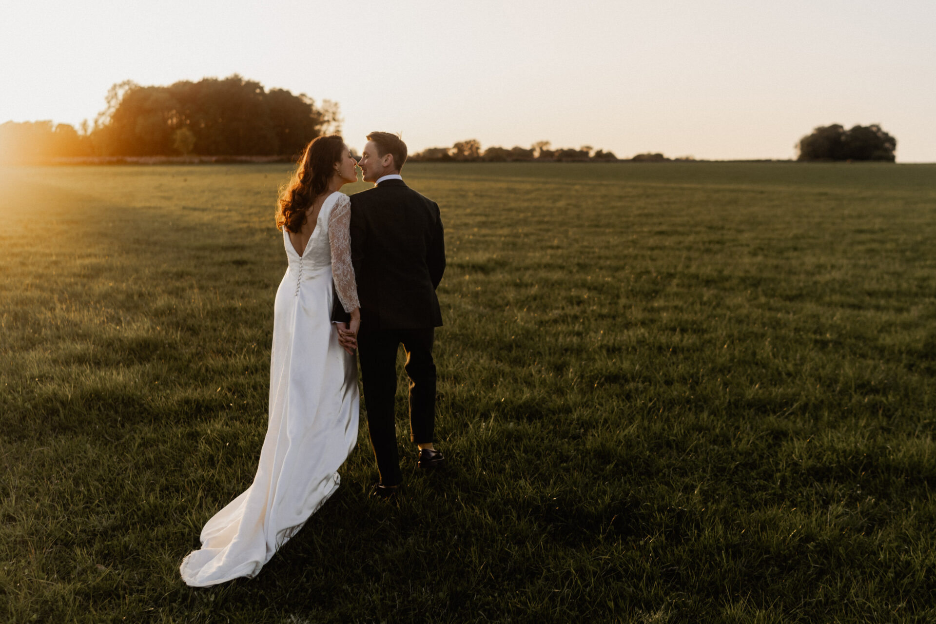 A bride and groom walk hand-in-hand across a green field at sunset. The bride is in a long white dress, and the groom is in a dark suit. This moment from their 2023 wedding photos will probably be one of their favorites.