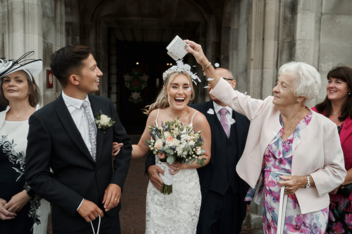 A happy bride playfully throws confetti around her groom outside a church, enjoying their wedding day to the fullest, with their excited guests around them.