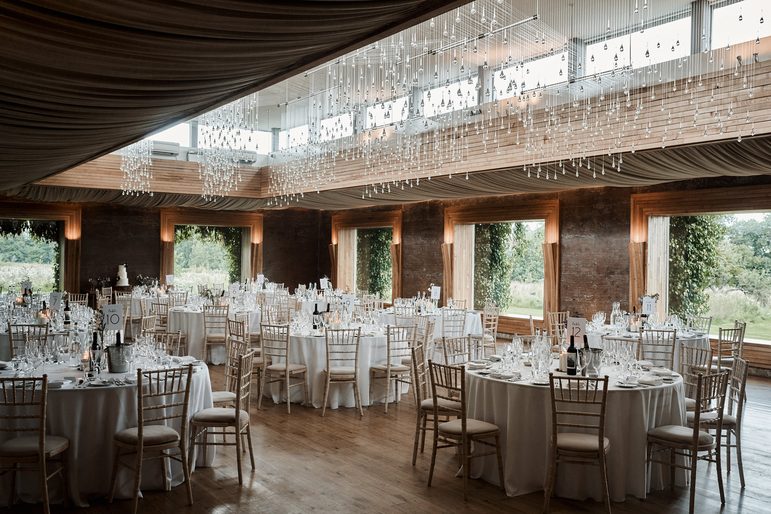 A big room with white tables and hanging lights for a wedding party.