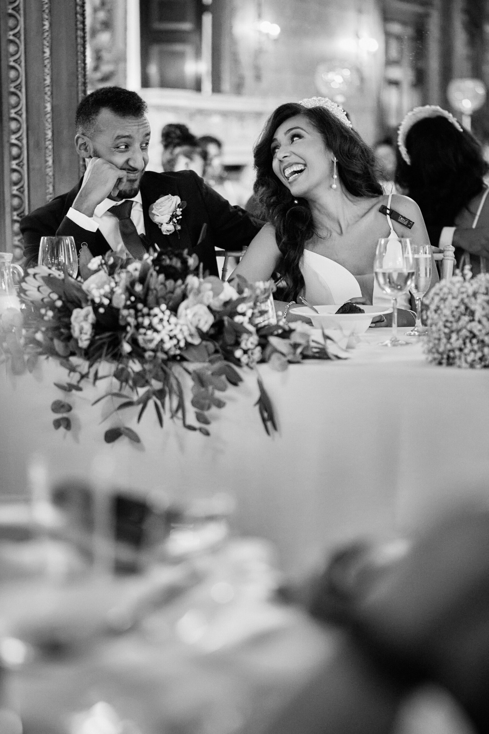 A black and white picture of a couple in their wedding attire sitting at a table.