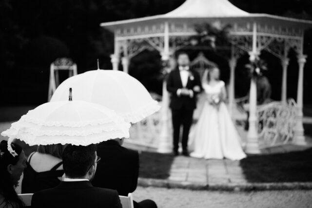 A man and woman getting married, holding umbrellas in front of a small outdoor structure.