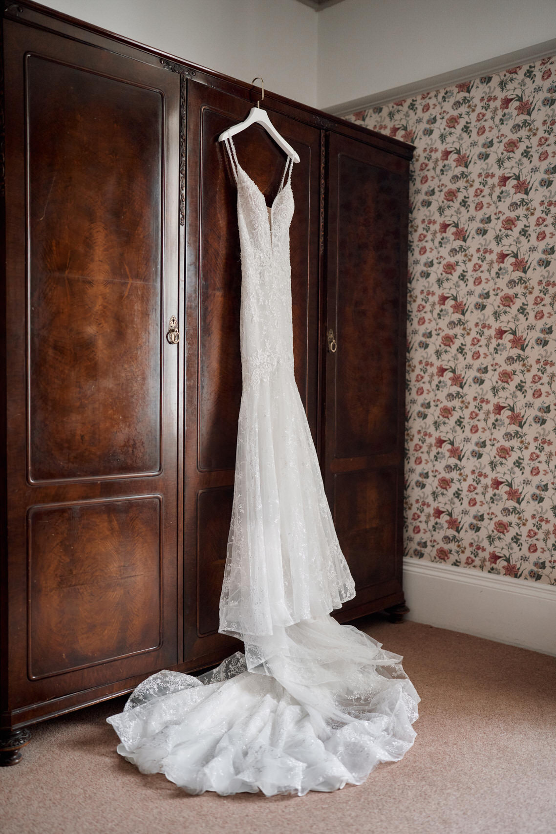 A wedding gown is hanging on a wardrobe in a room decorated with flower-patterned wallpaper.