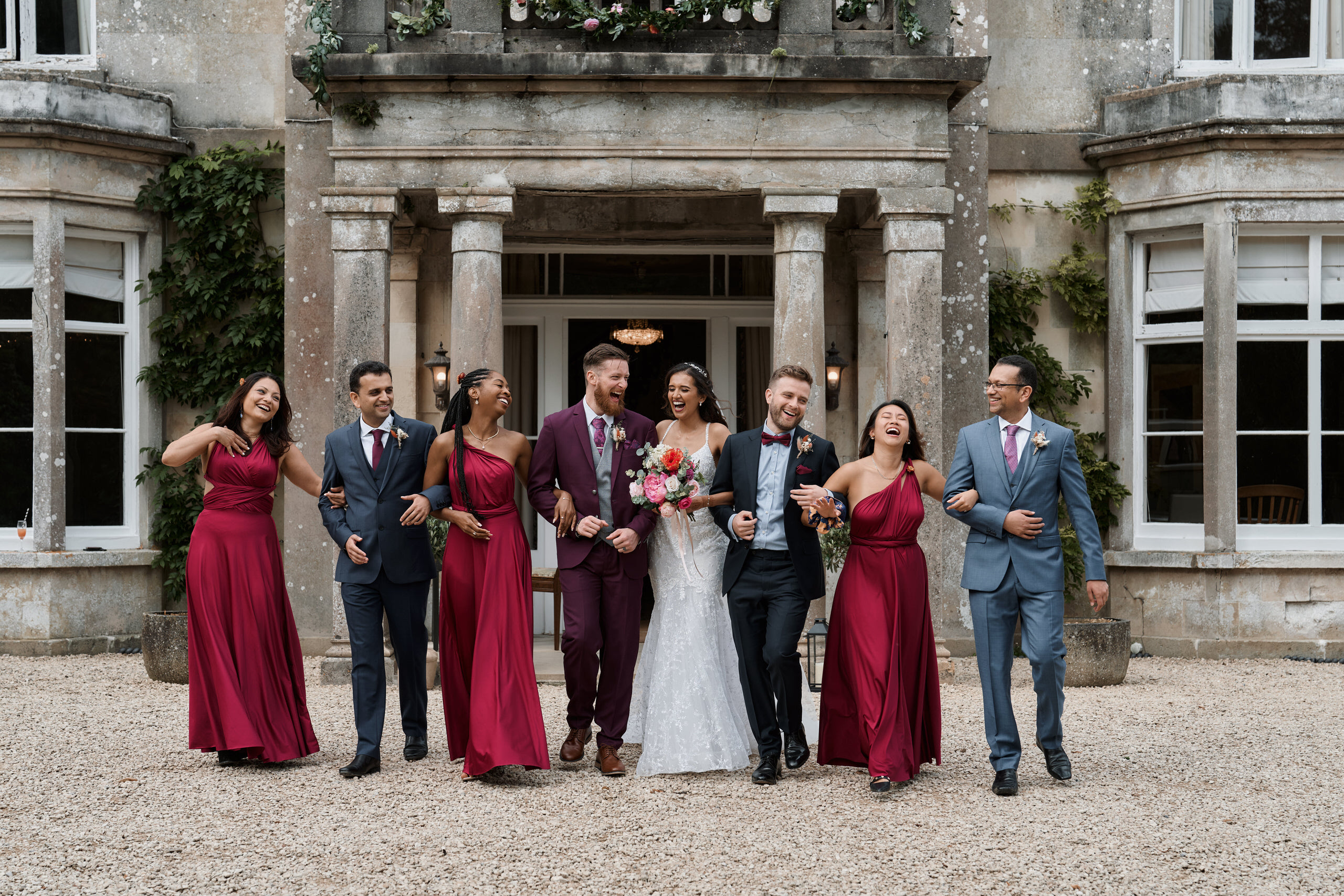 A bunch of bridesmaids and groomsmen are striking a pose in front of a big fancy house.