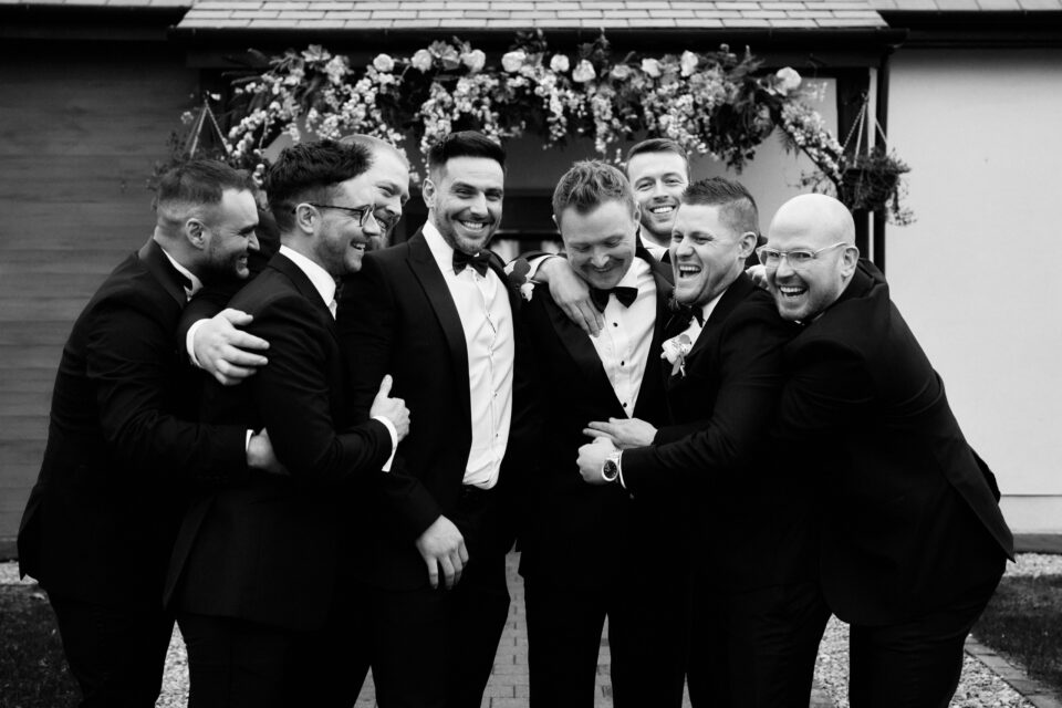 A black and white picture showing a bunch of guys at a wedding giving each other a hug.