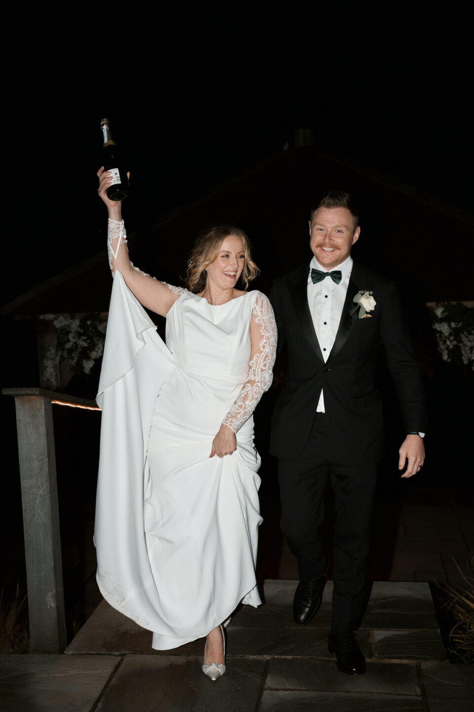 A couple getting married is holding a bottle of wine at night.