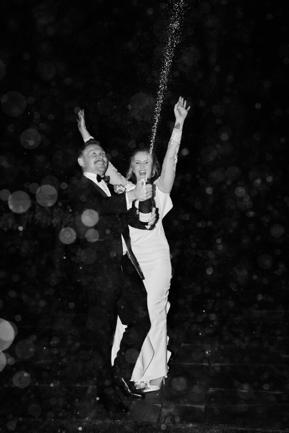 A picture in black and white showing a newlywed couple popping champagne.