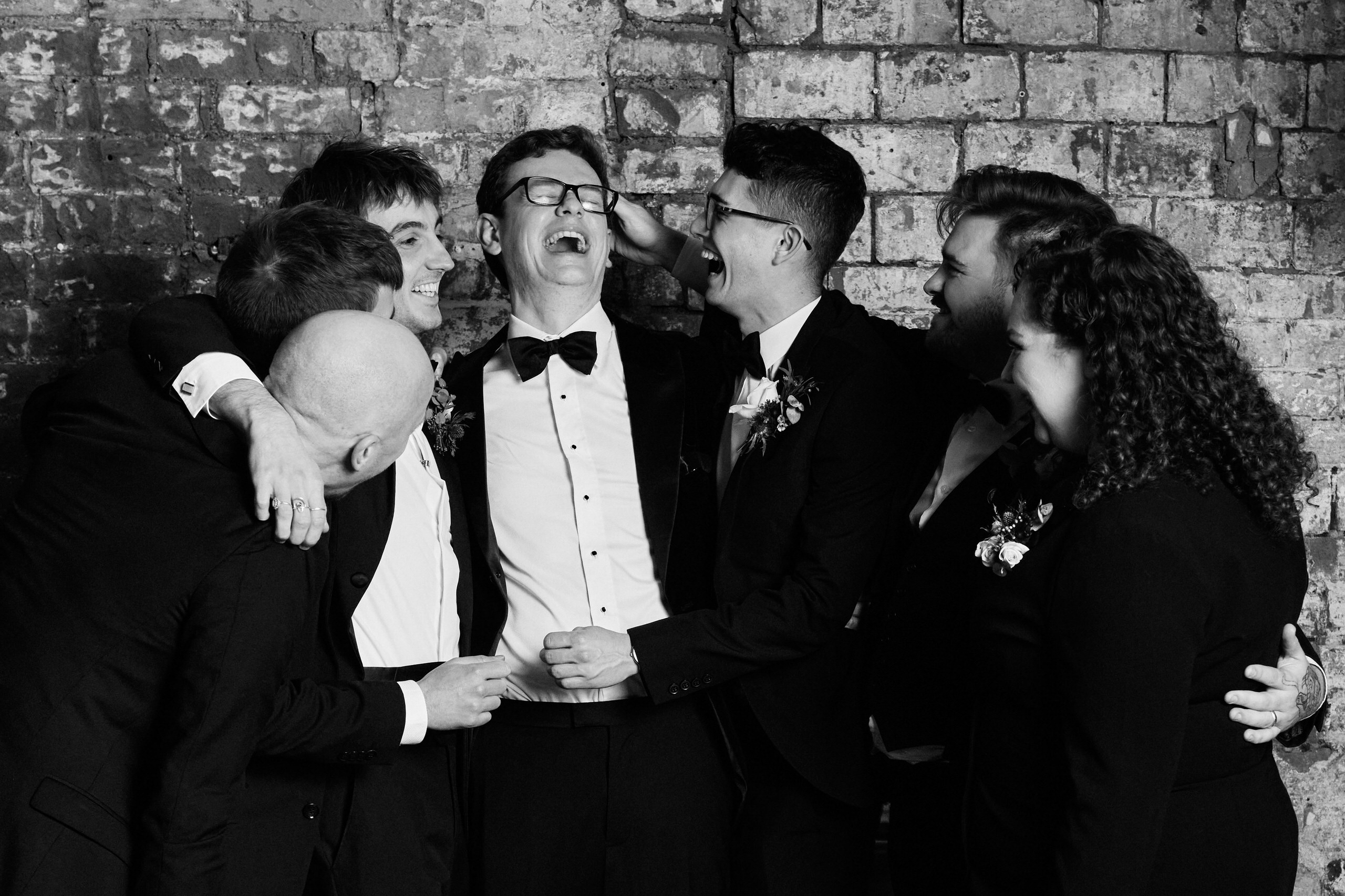 A bunch of groomsmen are hugging each other in front of a brick wall.
