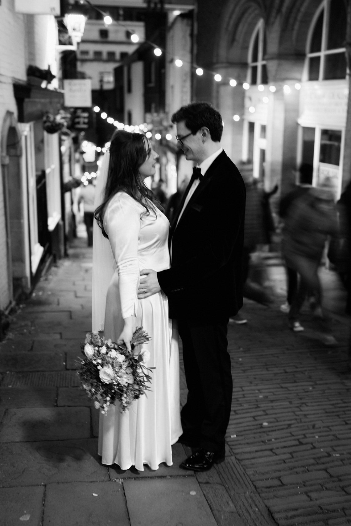 A couple just married standing on a stone-paved street at night.