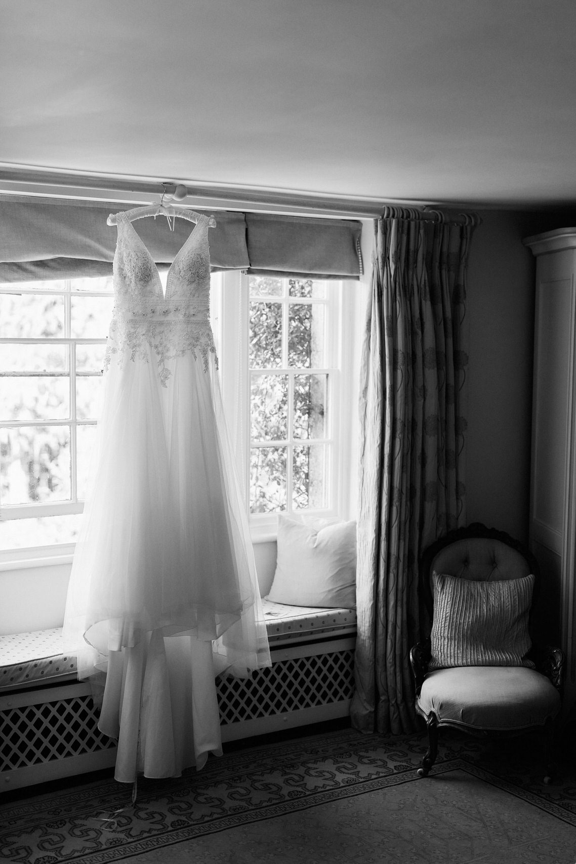 A photo in black and white showing a wedding dress hanging up near a window.