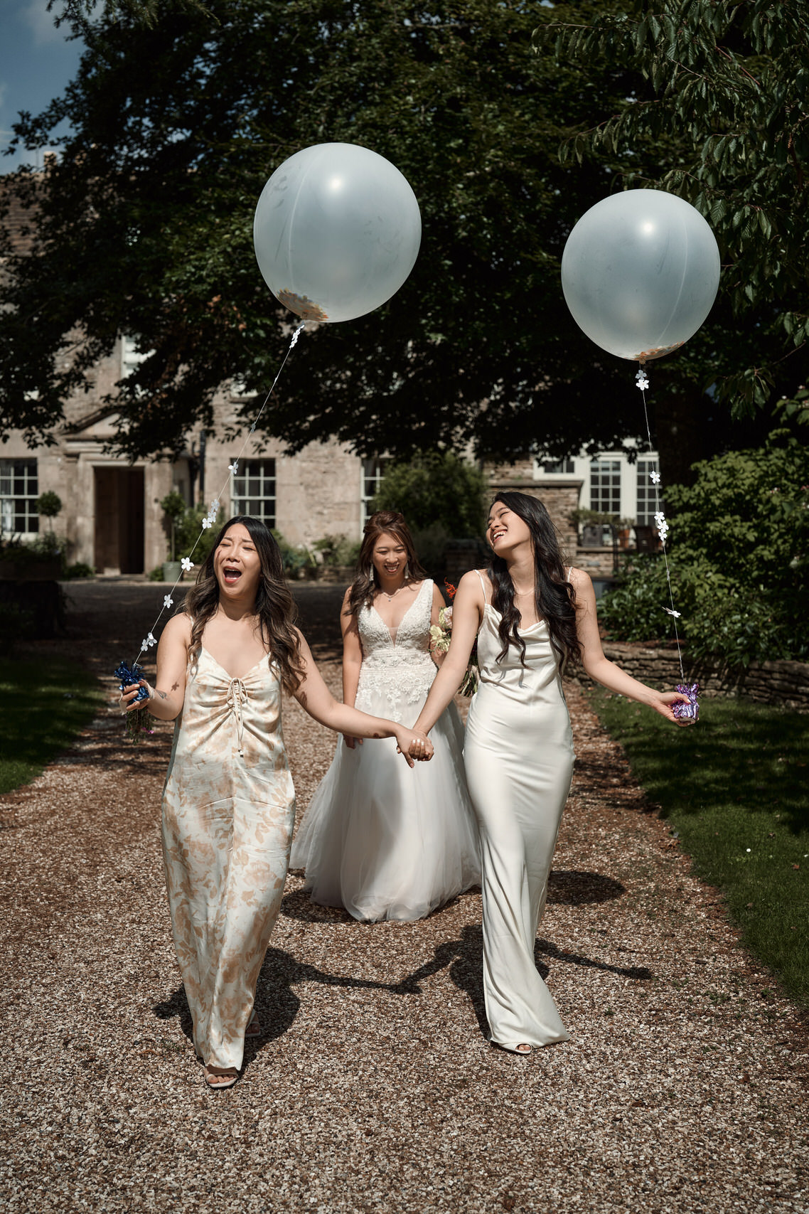 Three girls from a wedding are strolling down a pathway, carrying balloons.