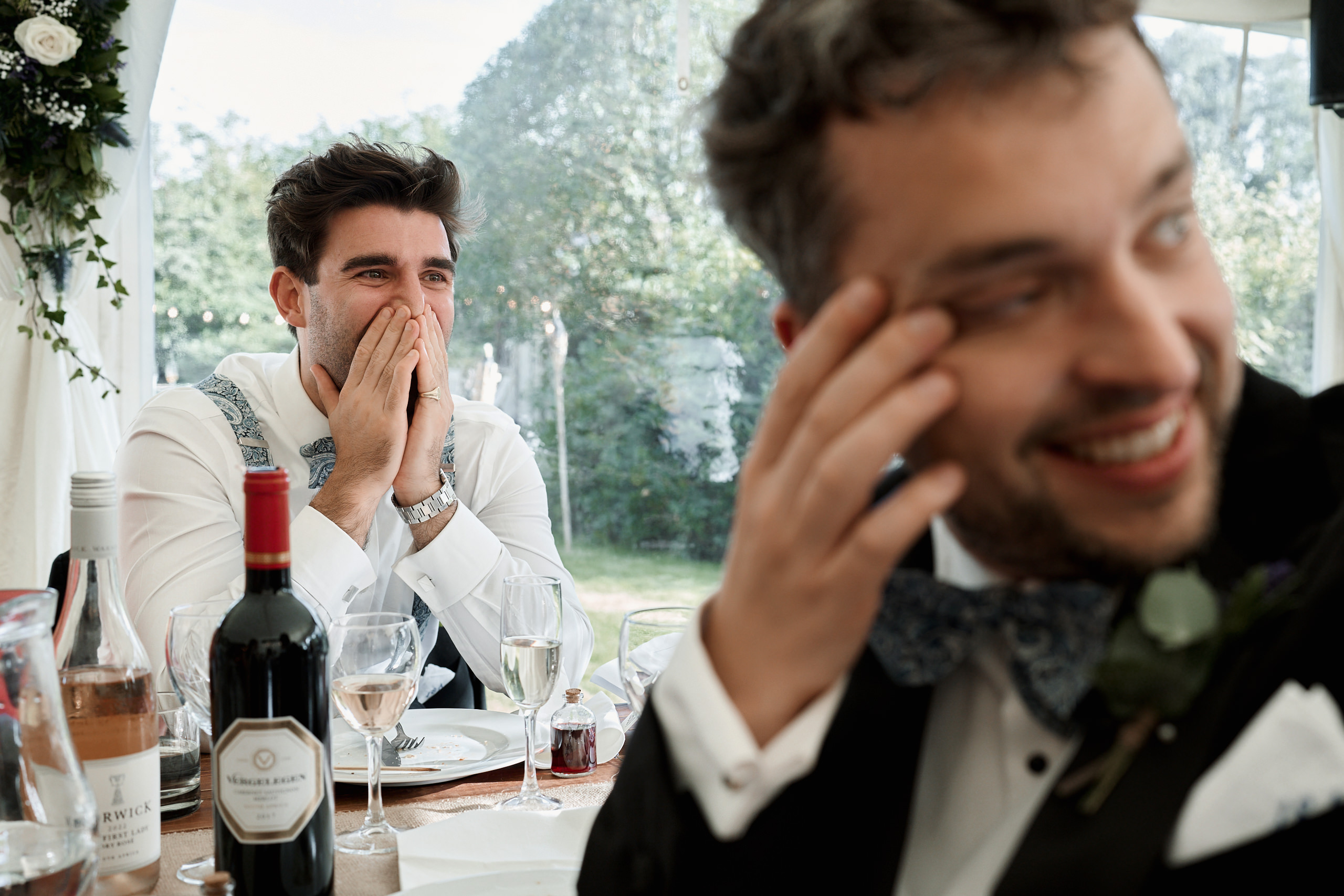 A guy in a tux and bow tie is sitting at a table with a bottle of wine.